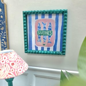 Puede incluir: Obra de arte enmarcada con un marco turquesa. Representa un rectángulo rosa con tres peces azules y la palabra "SARDINES" en una pancarta verde. El fondo tiene rayas azules y blancas.