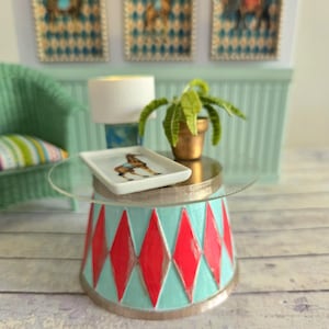 Op de afbeelding: Een miniatuur glazen tafel met een turquoise en rode basis met ruitvormig patroon. De tafel heeft een wit keramisch dienblad met een geprinte afbeelding van een giraffe. Een kleine plant in een pot staat op de tafel.