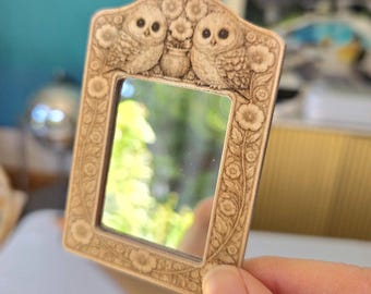May include: A small, rectangular mirror with a decorative wooden frame. The frame features carved details of two owls, flowers, and foliage. The mirror is held by a hand, and the background is blurred.