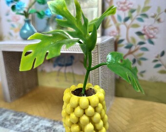 May include: A miniature yellow pineapple-shaped planter with a green plant. The planter is filled with soil and has a textured surface. The plant has large, green leaves. The background includes a mirror and floral wallpaper.
