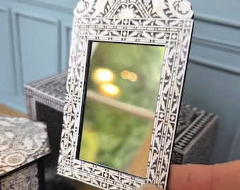 May include: A miniature decorative mirror with an ornate white and black frame. The mirror has a rectangular shape and is held by a hand. The frame features detailed patterns and designs, adding a touch of elegance. The mirror reflects a blurred outdoor scene.