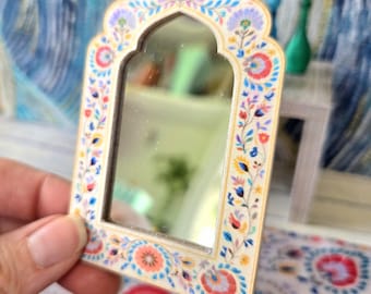 May include: A small, arched mirror featuring a cream-colored frame decorated with a colorful floral pattern in shades of pink, blue, yellow, and green. The mirror has a pointed, decorative top edge and a reflective glass center surface.