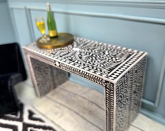 May include: A black and white patterned console table with a gold tray, champagne bottle, and a glass of champagne. The table has a decorative floral design and is placed in front of a light blue wall.
