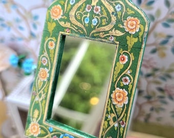 May include: A small, ornate mirror with a green frame. The frame is decorated with gold accents and colorful floral patterns in orange, pink, and blue. The mirror has a rectangular shape with a curved top.