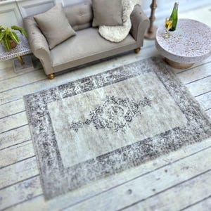Puede incluir: Una alfombra gris claro con un intrincado patrón floral gris oscuro. La alfombra tiene un aspecto desgastado y está colocada sobre un suelo de madera clara.