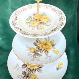 May include: Three-tiered serving stand with a gold handle. Each tier features a white porcelain plate with a gold rim and a floral design of yellow roses and brown leaves.