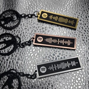 May include: Three metal keychains with Spotify code designs. One is gold, one is rose gold, and one is silver. Each keychain has a black chain and a Spotify logo. The keychains are rectangular with a barcode-like pattern.