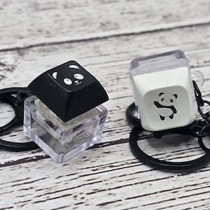 May include: Two keychains with panda designs. One keychain has a black keycap with a white panda face. The other keychain has a white keycap with a black panda illustration. Both keycaps are attached to clear plastic cases and black key rings.