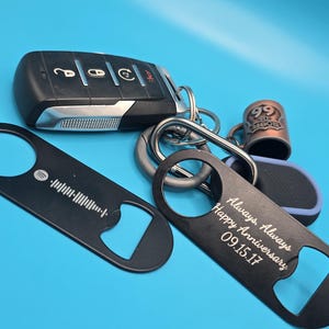 Puede incluir: Una colección de llaveros y accesorios sobre un fondo azul. Incluye una llave de coche, dos abridores de botellas negros, uno con un código Spotify y el otro con el texto "Always, Always Happy Anniversary 09.15.17", y un llavero de la Ruta 66.