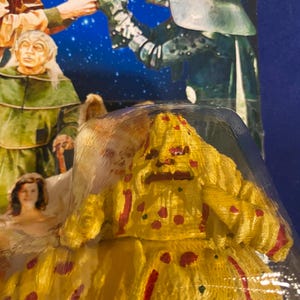 May include: A yellow action figure with red spots and streaks, resembling a character from a science fiction film. The figure is in a plastic package, with a background featuring other characters and a starry sky.