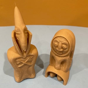 May include: Two 3D printed figurines, one with a long, pointed nose and the other with a round head and a wide, open mouth. Both figurines are beige in color and have a textured surface.