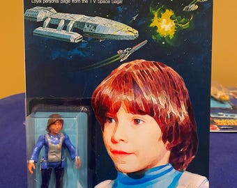 Può includere: Una action figure vintage di Boxey della serie televisiva Battlestar Galactica. La figura indossa una tuta blu e si trova su uno sfondo blu. La figura è confezionata in una confezione blister di plastica con un retro in cartone. Il retro presenta un'immagine della nave spaziale Battlestar Galactica e il testo "Battlestar Galactica" e "Boxey".