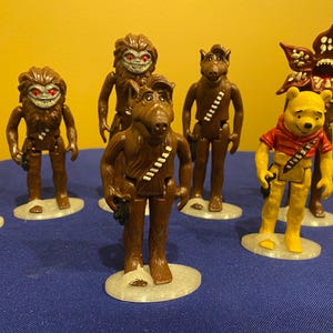 May include: A collection of brown action figures, including a bear-like character in a red shirt, and several other figures with unique designs. The figures are standing on small, light-colored circular bases, set against a blue surface.