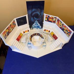 May include: A white plastic science fiction command center playset featuring a central circular console, multiple white chairs, and tiered platforms. The walls are decorated with colorful geometric grid patterns and a tall central space-themed backdrop.