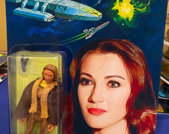 Può includere: Action figure di Battlestar Galactica Serina su una carta blu e nera. La carta presenta una scena spaziale con un'astronave e il testo "News Reported turned Viper Pilot in the TV Space Saga!". La figura è in una custodia di plastica trasparente.