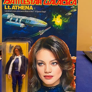 May include: Battlestar Galactica Lt. Athena action figure in original packaging. The packaging is blue and yellow with images of a spaceship and the character. The figure is wearing a blue uniform with a white trim.