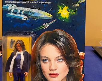 Può includere: Action figure di Battlestar Galactica Lt. Athena nella confezione originale. La confezione è blu e gialla con immagini di un'astronave e del personaggio. La figura indossa un'uniforme blu con finiture bianche.