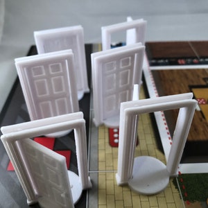 May include: Five white plastic doors with a raised design, standing on a game board with red and white stripes. The doors are different sizes and some are open.