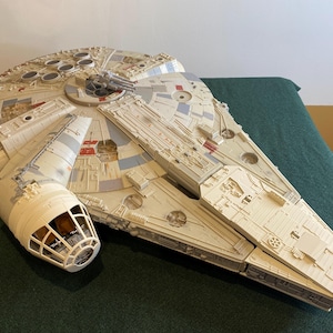 Puede incluir: Un juguete de nave espacial Millennium Falcon blanco y gris con detalles rojos. El juguete tiene una cabina y un tren de aterrizaje desmontable.
