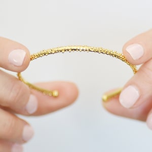 May include: A gold bangle bracelet with a textured surface. The bracelet is made of small, round beads.