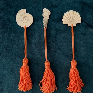 Puede incluir: Tres abalorios de marcapáginas de plata con borlas naranjas. Los abalorios tienen forma de concha, caracol y nautilus.