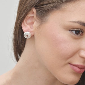 Puede incluir: Pendientes de botón de plata con forma de erizo de mar. Los pendientes son pequeños y delicados, y tienen una superficie texturizada.