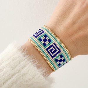May include: A wide beaded bracelet with a geometric pattern in shades of blue, teal, and white. The bracelet has a gold-colored border. The design features repeating square and spiral motifs. The bracelet is worn on a wrist.