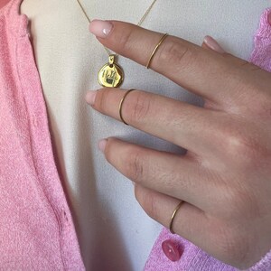Può includere: Una collana d'oro con un ciondolo rotondo su cui è scritto "Hello". La collana è indossata da una persona con un cardigan rosa e anelli d'oro alle dita.
