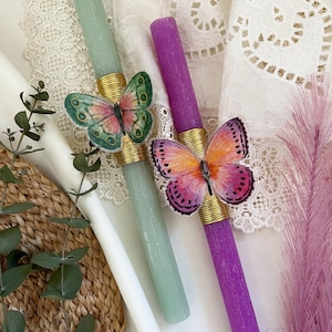 Puede incluir: Dos velas cónicas, una verde claro y otra morada, adornadas con decoraciones de mariposas. Las mariposas tienen alas rosas, naranjas y verdes. Bandas doradas rodean la base de cada mariposa y vela.