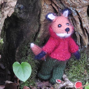 May include: A knitted fox toy wearing a red jumper and green trousers. The fox is standing in front of a tree trunk with moss growing on it.