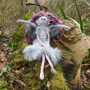 May include: A knitted fairy doll with purple yarn hair, gray wings, and a gray and purple dress. The doll is sitting on a mossy tree stump in a forest setting.