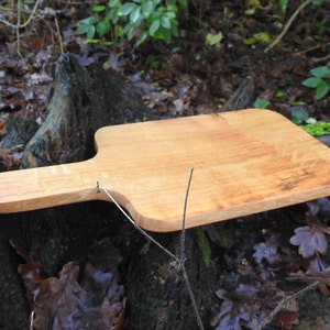 May include: A wooden cutting board with a handle, made of light brown wood. The board is sitting on a tree stump.