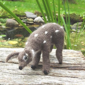 May include: A handmade knitted deer figurine in shades of brown with white spots. The deer is posed with its head down, showcasing its delicate features. The figurine is set against a natural outdoor backdrop, highlighting its handcrafted nature.