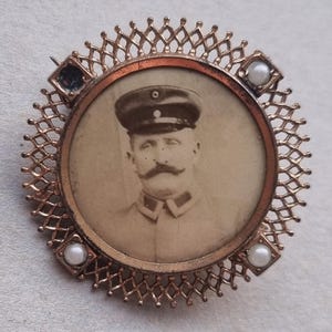 Peut inclure: Broche circulaire vintage avec un portrait sépia d'un homme en uniforme militaire. Le portrait est encadré par un filigrane en métal couleur cuivre, orné de petites perles et de pierres carrées de couleur foncée. La broche a une épingle au dos.