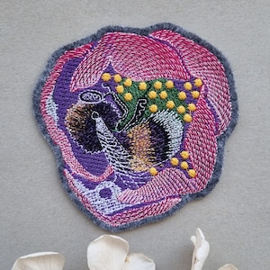 Può includere: Un cerotto ricamato a forma di fiore rosa e viola con accenti gialli e verdi. Il cerotto è su uno sfondo grigio.