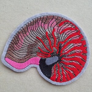 Könnte beinhalten: Gestickter Aufnäher in Form einer Nautilusmuschel. Der Aufnäher hat einen grauen Rand, wobei das Muscheldesign in Rosa-, Braun-, Schwarz- und Rottönen gehalten ist. Das Design ist mit aufwendigen Stichen detailliert, wodurch ein strukturiertes Aussehen entsteht.