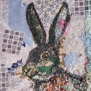 May include: A close-up of a black and white embroidered image of a rabbit's head on a pink and white fabric background. The rabbit's fur is detailed with black thread and the background is made of various patterned fabrics.