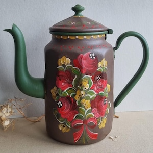 Könnte beinhalten: Eine braune Metallteekanne mit grünem Griff, Ausguss und Deckel. Der Körper ist mit einem handgemalten Blumenmuster aus roten Rosen, gelben Akzenten und einer roten Schleife verziert. Der Deckel hat einen grünen Knauf und weiße Punkte.