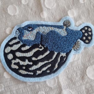 Könnte beinhalten: Ein blau-weiß gestickter Kugelfisch-Aufnäher mit hellblauem Rand. Der Fisch hat detaillierte Stickereien, einen schwarz-weiß gestreiften Bauch und einen strukturierten blauen Körper. Geeignet für Kleidung oder Accessoires.