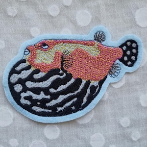 Könnte beinhalten: Gestickter Aufnäher mit einem Kugelfisch-Design. Der Fisch hat einen rot-gelben Körper mit schwarz-weißen Details. Der Aufnäher ist hellblau umrandet.