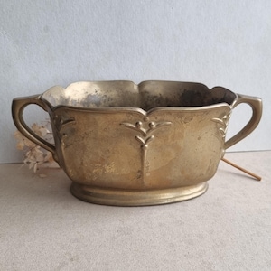Könnte beinhalten: Eine ovale Messingschale mit zwei seitlichen Griffen und einem gewellten Rand. Die Außenseite weist ein erhabenes, symmetrisches, botanisch inspiriertes Reliefmuster auf. Die Metalloberfläche zeigt eine gealterte Patina mit Flecken.