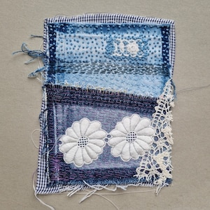 Może przedstawiać: Niebiesko-biała tkanina patchworkowa z dwoma białymi haftowanymi kwiatami i kratkowanym obramowaniem.