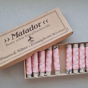 May include: A vintage box of "Matador" buttonhole thread, with the text "Beste echte Knopflochseide". The box is open, revealing several spools of light pink thread. The box is a light brown colour, and the spools are on a light brown cardboard base.