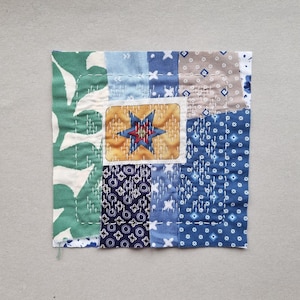 Può includere: Un tessuto patchwork quadrato con vari modelli e colori di tessuto. Il design include stampe di foglie verdi, forme geometriche blu e bianche e un rettangolo centrale con un motivo a stella. I dettagli cuciti a mano sono visibili.