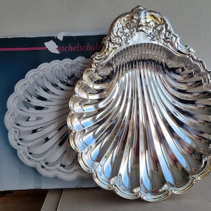 Könnte beinhalten: Eine polierte silberfarbene Metallschale in Form einer Jakobsmuschel mit detaillierten floralen Prägungen am oberen Rand. Die Schale weist tiefe, strahlenförmige Rillen auf und ist vor ihrer originalen Kartonverpackung positioniert.