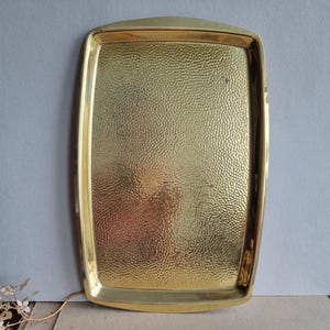 Könnte beinhalten: Ein rechteckiges, goldfarbenes Metalltablett mit einer gehämmerten Textur auf der Oberfläche. Das Tablett hat glatte, erhöhte Ränder und eine polierte Oberfläche, die das Licht reflektiert und einen hellen Glanz auf dem Bereich erzeugt.