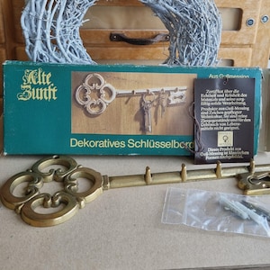 Könnte beinhalten: Ein Schlüsselbrett aus Messing in Form eines Schlüssels mit vier Haken, verziertem Kopf und einem Ende in Schlüssellochform. Das Produkt wird mit der originalen grünen Kartonverpackung geliefert, die den Text Alte Zunft und Dekoratives Schlüsselbord zeigt.