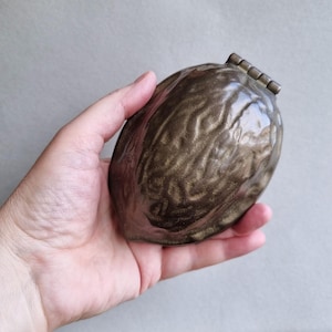 Op de afbeelding: Een bronzen, walnootvormige sieradendoos met een scharnierend deksel. De doos heeft een textuur, die lijkt op de natuurlijke ribbels van een walnootschil. De doos wordt in een hand gehouden tegen een neutrale achtergrond.