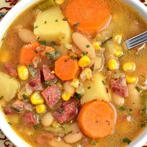 Puede incluir: Un cuenco de sopa abundante con zanahorias, patatas, maíz y frijoles. La sopa tiene un caldo claro y se sirve en un cuenco blanco sobre un plato decorativo. Una ramita de romero es visible en el fondo.