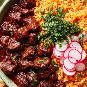 May include: A bowl of Mexican food with braised meat in a rich sauce, orange rice, fresh cilantro, and sliced radishes. The dish is served in a green bowl, with lime wedges and tortillas in the background.